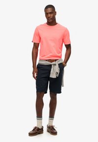 Herreoutfit bestående af en lys pink bomulds t-shirt, marineblå shorts, en grå sweater bundet om livet, og brune sko med mønstrede sokker.