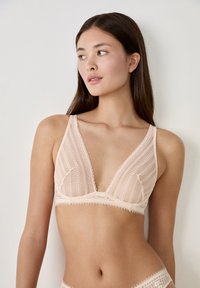 Lichtroze kant bralette met een diepe V-halslijn, zachte driehoekige cups en delicate gegolfde randen, die het sheer, gestructureerde stof benadrukken.