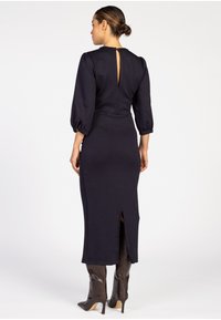 Marineblaues langärmeliges Kleid mit einem Rückenschlitz, figurbetontem Design und Schlitz am Saum; kombiniert mit kniehohen, glänzenden braunen Stiefeln.