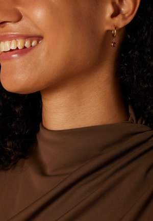 Gouden hoepel oorbel met een kleine rode edelsteen, gedragen aan het oor. Gladde huid en textiel met een bruine textuur gedrapeerd rond de nek.