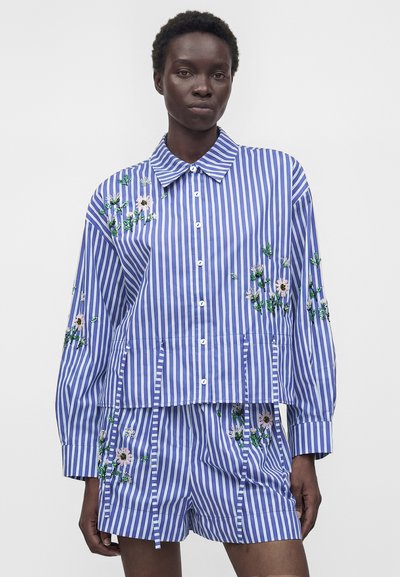 Femme noire portant une chemise à rayures verticales bleu et blanc à manches longues et un short avec broderie florale.