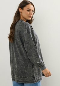 Felpa grigia oversized con lavaggio acido, maniche lunghe, polsini a costine e orlo sventagliato, esposta su uno sfondo neutro.