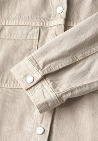 Beige shirt jacket made of textured fabric, featuring silver buttons and a visible seam. Cuffs and pockets are also detailed in the fabric.