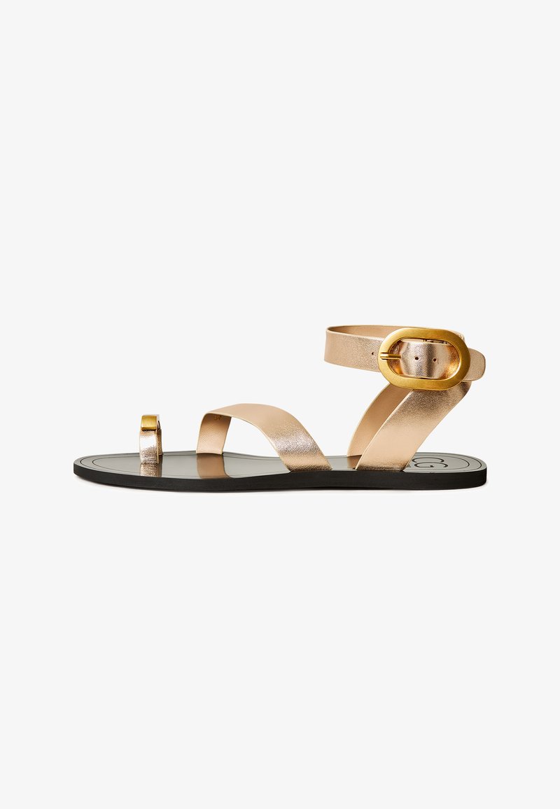 Sandales en or métallique avec plusieurs lanières, un bout carré et une boucle dorée proéminente. La semelle est noire avec un design plat.