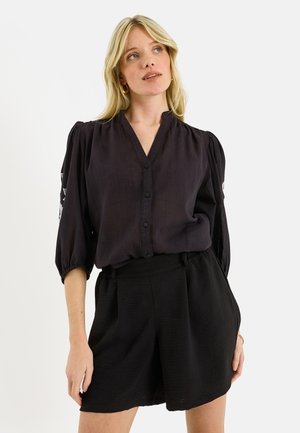 Mujer con cabello rubio que lleva una blusa negra abotonada con mangas abullonadas y pantalones cortos negros, de pie frente a un fondo claro y liso.