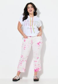 Vit kortärmad huvtröja med färgglad text och snören, ihop med korta vita jeans med rosa splattermönster och svarta sandaler.