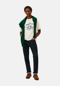 Hombre con camiseta gráfica blanca, jeans oscuros, zapatos marrones y un suéter de punto verde colgado sobre los hombros, de pie con las manos en los bolsillos.