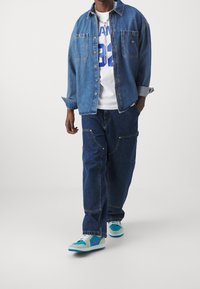 Kurtka dżinsowa z dwoma kieszeniami na piersi, nałożona na białą koszulkę z niebieskim napisem. Luźne dżinsy i wielokolorowe sneakersy.