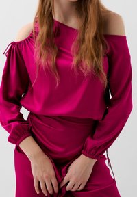 Haut en soie fuchsia avec design épaules dénudées, manches longues bouffantes et détails froncés aux épaules. Texture lisse avec une coupe ample.