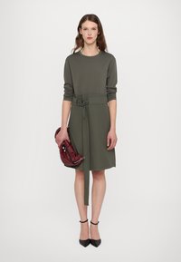 Abito verde oliva con maniche lunghe, vita con cintura e orlo a lunghezza ginocchio. La modella tiene una borsa bordeaux e indossa scarpe col tacco nere.