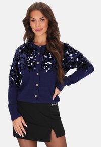 Donna che indossa un cardigan blu navy con paillettes e bottoni e una mini gonna nera con uno spacco frontale, in posa con una mano sull'anca.