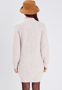 Vestito maglia beige chiaro con colletto alto e maniche lunghe, caratterizzato da una texture a costine e una vestibilità morbida che arriva sopra le ginocchia.