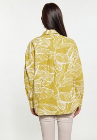 Camicia gialla con motivo a foglia bianca, maniche lunghe, vestibilità rilassata e dettaglio sul retro. Indossata con pantaloni chiari.
