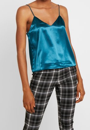 Kobieta ubrana w satynowy turkusowy top na cienkich ramiączkach, w połączeniu z dopasowanymi czarno-białymi spodniami w kratę, stojąca na tle jasnoszarego tła.