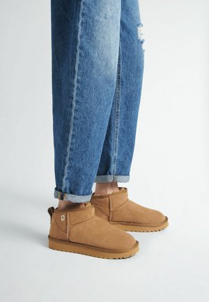 Stivaletti in suede marrone con punta rotonda, dettagli cuciti e suola piatta in gomma, abbinati a jeans in denim blu con risvolti.