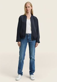 Tamsiai mėlyna jūreivio stiliaus bombonešio striukė su ribinėmis rankogaliais, dėvima virš balto marškinėlio, derinama su mėlynomis džinsais ir baltais sportbačiais. Lygus audinys ir laisvas, kasdienis modelis.