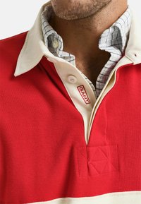 Rode poloshirt met een witte kraag, voorzien van een horizontale witte baan. Bevat gestructureerde accenten en een logo label in het rood.