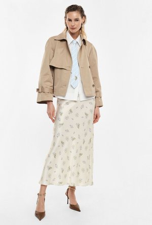 Donna che indossa un giubbotto beige corto, camicia bianca con cravatta azzurro chiaro, gonna lunga crema a fiori e tacchi alti marroni, in piedi davanti a uno sfondo bianco.