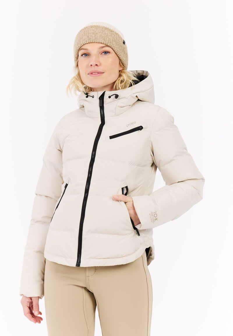 Weiße gepolsterte Jacke mit Kapuze, schwarzen Reißverschlüssen, elastischen Bündchen und einer Reißverschlusstasche auf der Brust. Trägt eine beigen Strickmütze.