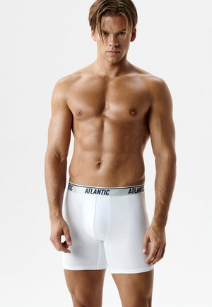 Mężczyzna o umięśnionej sylwetce, ubrany w białe bokserki marki Atlantic, stojący na tle jednolitego białego tła, zwrócony przodem.
