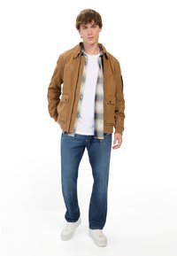Brun ruskindsjakke med klaplommer, lagdelt over en ternet skjorte, hvid T-shirt, blå jeans og hvide sneakers. Afslappet pasform, ensfarvet baggrund.