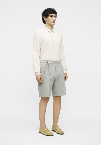 Άνδρας με σκούρα σγουρά μαλλιά, φορώντας μια κρεμ πουκαμίσα με μακριά μανίκια και κουμπιά, ανοιχτό γκρι σορτς με ζώνη και μπεζ μοκασίνια, στέκεται μπροστά σε λευκό φόντο.