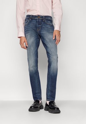 Mand iført slim-fit falmede blå jeans, lyserød skjorte med knapper og sorte læder loafers, stående mod en ensfarvet baggrund.
