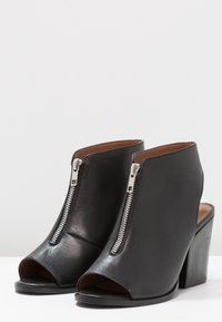 Czarne skórzane botki za kostkę z otwartym palcem i srebrnymi suwakami z przodu, na chunky obcasach, na białej powierzchni.