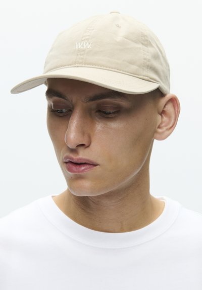 Νέος άνδρας που φοράει μπεζ καπέλο με λευκό κείμενο "www.", κοιτάζει προς τα κάτω και προς το πλάι, ντυμένος με λευκό μπλουζάκι με στρογγυλή λαιμόκοψη.