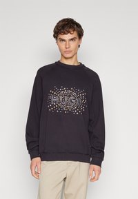 Sudadera negra con cuello y puños de canalé, que presenta un patrón de tachuelas metálicas que forman la palabra "HUGO" en un diseño circular.
