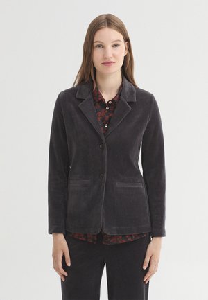 Young woman wearing a dark gray corduroy blazer over a patterned shirt, standing against a plain light background.