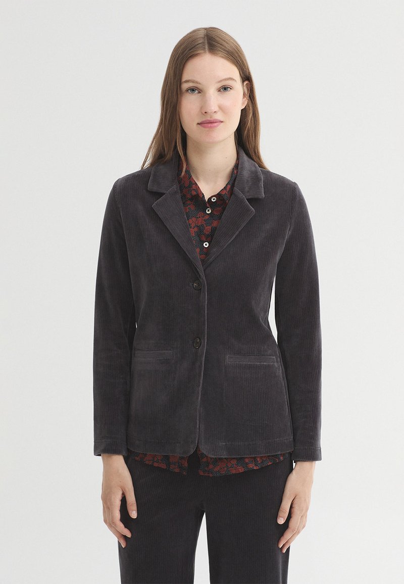 Jeune femme portant un blazer en velours côtelé gris foncé par-dessus une chemise à motifs, debout devant un fond clair uni.
