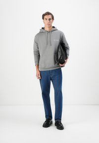 Grå hettegenser med frontlomme, kombinert med blå denimjeans. Modellen holder en svart veske laget av glatt, teksturert materiale.