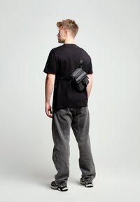 Svart crossbody-väska i texturerat tyg, med flera blixtlåsfack, bärs över en enkel svart T-shirt och urblekta gråjeans.