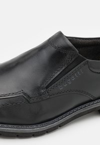 Zapato negro de cuero tipo slip-on con paneles laterales elásticos, detalles de costura y suela de goma texturizada. Nombre de la marca "bugatti" grabado en el lado.