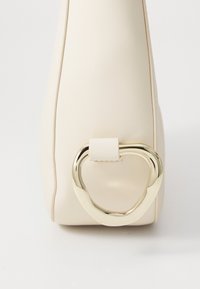 Cream faux leather handbag with a smooth texture, featuring a gold heart-shaped metal accent and a small strap attachment.