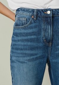 Vaqueros de denim azul con cintura alta, que cuentan con un diseño tradicional de cinco bolsillos, remaches de cobre y detalles de costura en contraste.