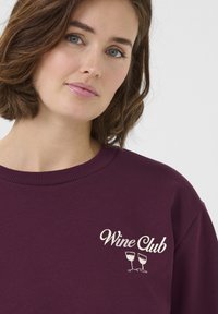 Sweatshirt bourgogne avec le texte blanc "Wine Club" et un graphique de deux verres à vin ; col côtelé et texture douce.
