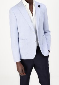 Světle modrý blazer s texturovaným povrchem, jednoradový design, dvě přední kapsy a dekorativní černá květinová brož na klopě.