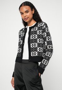 Melna un balta cropped kardigāna ar ģeometriskiem rakstiem, teksturētu audumu un pogu aizdari. Valkā virs baltas krekla un melnām biksēm.