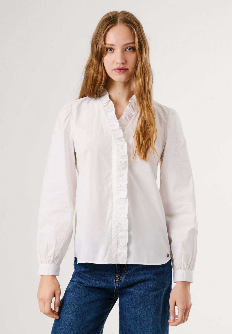 Giovane donna con lunghi capelli ondulati, che indossa una blusa bianca a balze e jeans blu, in piedi contro uno sfondo chiaro e semplice.