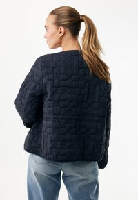 Granatowa pikowana kurtka o fakturowanym wzorze, z okrągłym dekoltem i luźnym krojem, zestawiona z jasnoniebieskimi jeansami.
