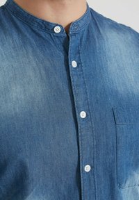 Homme portant une chemise en denim bleu avec un col chinois, des boutons blancs et une poche poitrine.