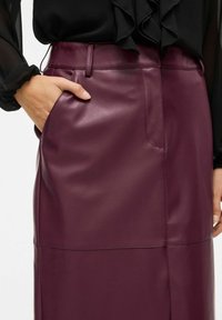 Ruka ženy v vrecku vysokého pásu, tmavo fialovej koženej sukne, skombinovanej s čiernym blúzkou s volánikmi.