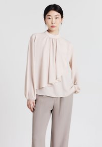 Blusa beige chiaro con dettagli plissettati, collo alto e maniche lunghe. Il tessuto appare liscio e morbido, in abbinamento a pantaloni grigi dal fit rilassato.