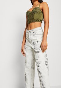 Olivgrön kort topp med spetsöverdrag och justerbara axelremmar, ihop med högmidjade, ljusgrå denimjeans med svarta streck.