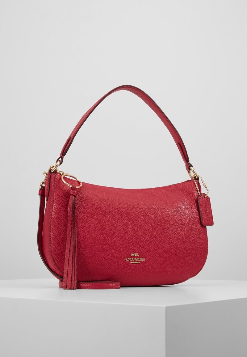 Sac à main en cuir rouge avec une texture granuleuse, forme semi-circulaire, poignée courte, détail à pampilles et une petite étiquette de marque en accents dorés.