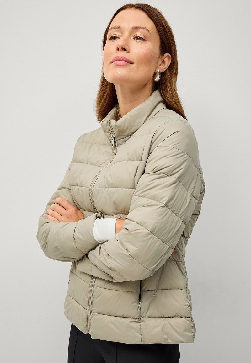 Chaqueta de plumas color beige claro con un cuello alto, diseño acolchado y cierre de cremallera. Textura suave con un ajuste relajado, combinada con una camiseta blanca underneath.