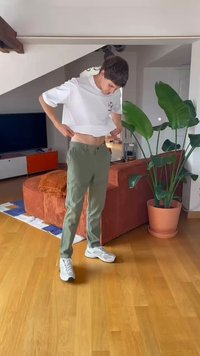 Witte cropped t-shirt en olijfgroene broeken. Casual sneakers. Binnenomgeving met een bruine bank en een plantenpot op de achtergrond.