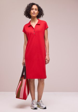 Kobieta z kręconymi włosami, ubrana w czerwoną sukienkę polo, srebrne trampki, trzymająca dużą paskowaną torbę w kolorach czerwonym, różowym i brązowym.